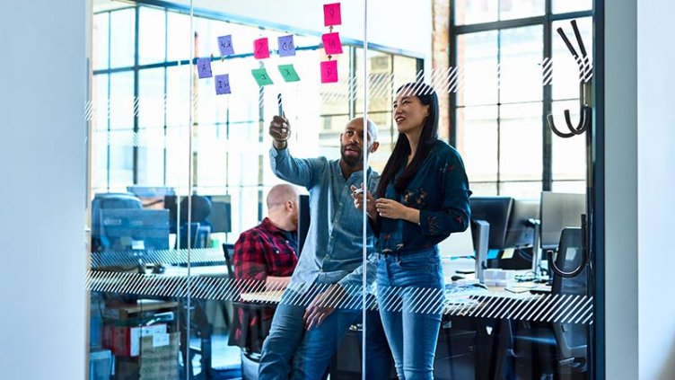 Two people in an office, positioned next to a wall filled with various sticky notes.